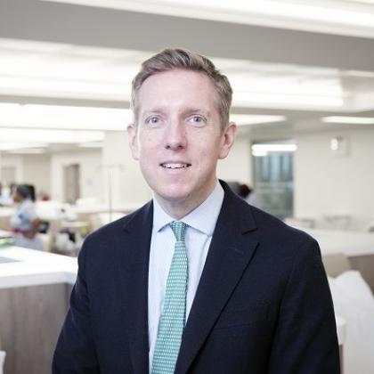 Head and shoulders of a man dressed smartly in a black suit jacket, white shirt and green patterned tie. He is smiling and sitting in a medical room in The Royal Marsden, with nurses in the background.