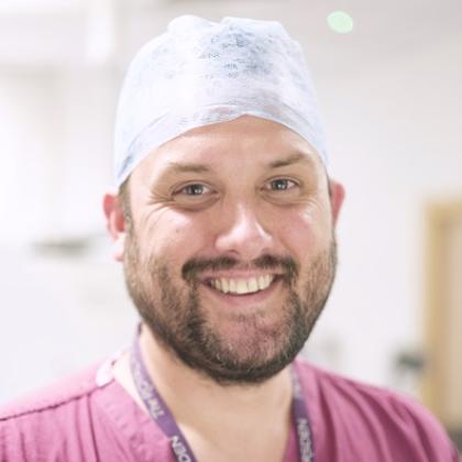 Headshot of man with beard wearing purple scrubs and white surgical cap, smiling