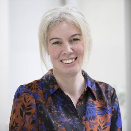 Headshot of Dr Minchom, she is wearing a smart, floral shirt and tanding in a bright room with large windows