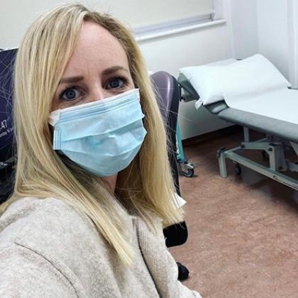 Head and shoulders of a lady with long blonde hair, wearing a facemask. Behind her is a hospital bed.