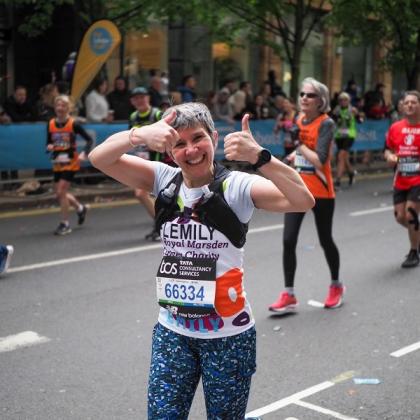 London Marathon runner, wearing a Royal Marsden Cancer Charity running vest. She's smiling and giving a double thumbs up at crowd as she jogs past.