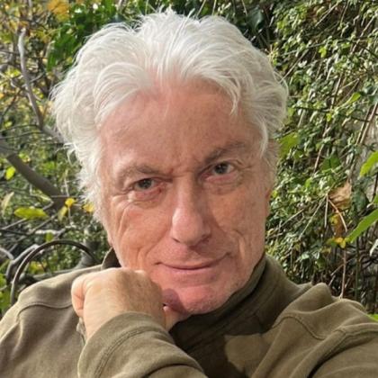 Headshot of Mike smiling outside in front of some shrubbery. He has silver hair and is wearing a brown jacket.