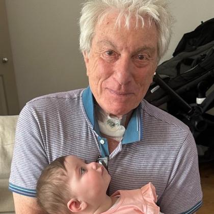 Man with a tracheostomy tube in his neck, smiling and holding a little baby on his lap. The baby is looking up at him.