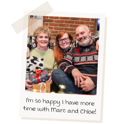 "I'm so happy to have more time with Marc and Chloe!". A polaroid of three people smiling with their arms round each other. Zoe, on the left, is wearing a green jumper and holding christmas presents, Chloe in the middle is wearing a patterned Christmas jumper. She has long auburn hair and is wearing glasses. Marc is on the right, he has a white beard and is wearing a Christmas jumper with reindeer on it.  