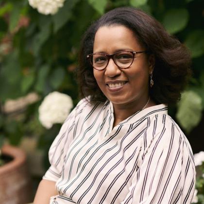 A Royal Marsden patient smiling and sitting in front of some potted flowers, wearing a stripped shirt and glasses with shoulder-length styled hair