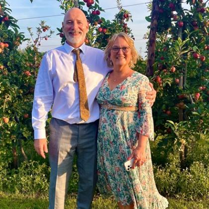 A man and a woman in smart clothes, standing smiling in front of fruit trees