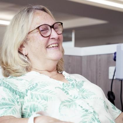 Upper body shot of woman with glasses and blonde hair, sitting next to hospital equipment, smiling