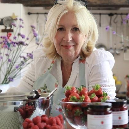 Fundraiser Sara who raises money for The Royal Marsden Cancer Charity selling jam. She is sitting in a kitchen, with purple flowers in the background, and kitchen utensils handing from the wall. She is wearing a fruit-patterned apron, white blouse and has had a bowl of strawberries in front of her and pots of jam.