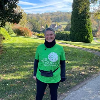 Woman in walking gear and charity t-shirt smiling. Trees are behind her and it's a sunny day