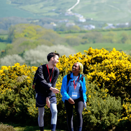 Two walkers smiling at each other, flowering bushes and hills behind them