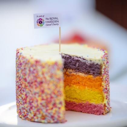 A tall cake with sprinkles on the outside and a buttercream icing on top. A slice has been taken out of the cake, revealing a middle of four layers of sponge, all different colours of pink, yellow, orange and purple. On top of the cake is a little flag topper that says 'In aid of The Royal Marsden Cancer Charity'.