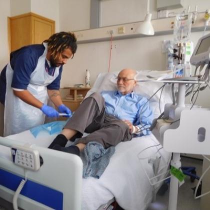 Richard laying on a hospital bed. A nurse in scrubs and an apron is lent over him and inserting a syringe into a drip attached to Richard.