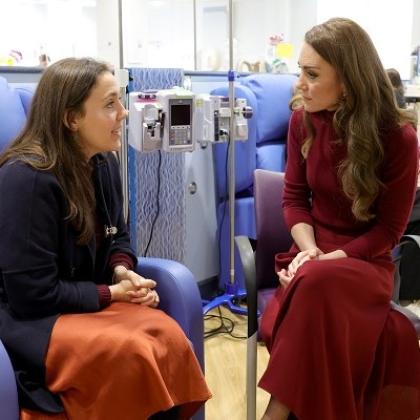 HRH The Princess of Wales sitting and listening intently to a young patient wearing a navy cardigan and orange silk dress, who is sitting in a treatment chair at The Royal Marsden.
