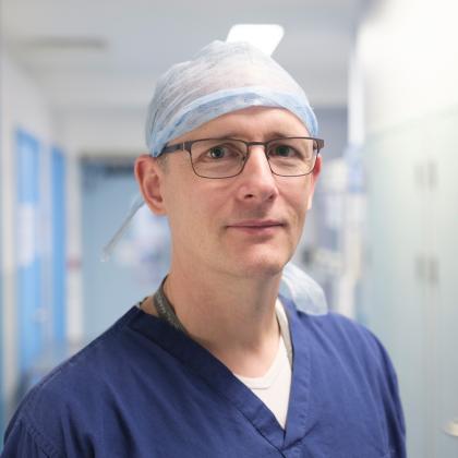 A man, Kieran Power, in dark blue medical scrubs, a blue cap and brown rimmed glasses