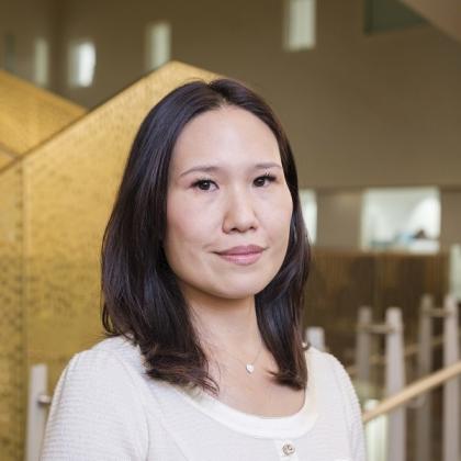 A headshot of Dr Irene Chong. She has dark, shoulder-length hair and is wearing a white top, paired with a white necklace. She is standing next to the golden staircase of the Oak Cancer Centre.