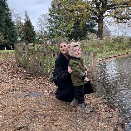 Tash crouched down with her young son, Rudi. They are next to a small pond in a countryside park with plenty of trees in the background. They are both wrapped up in coats and Rudi is wearing a fluffy hat with bear ears.
