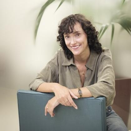 Royal Marsden patient, Nadia. She has dark curly hair and is wearing a smart, neutral coloured shirt with a gold necklace and watch. She's smiling and sitting on a chair, leaning on the backrest.