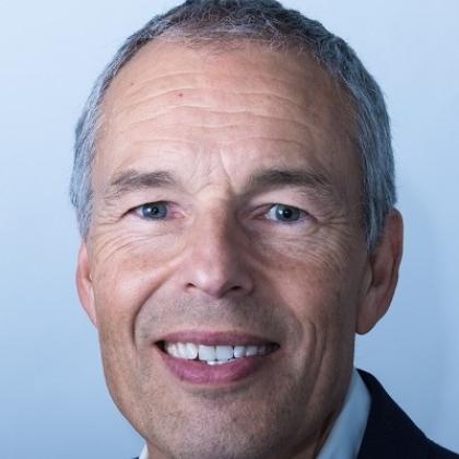 A headshot of Dennis. He is smiling, has short, silver hair and is wearing a suit.