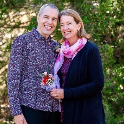 Dennis and his wife smiling and standing together in front of a tree. Dennis is wearing a smart, patterned shirt and his wife who is wearing a pink dress scar, with a long navy cardigan.