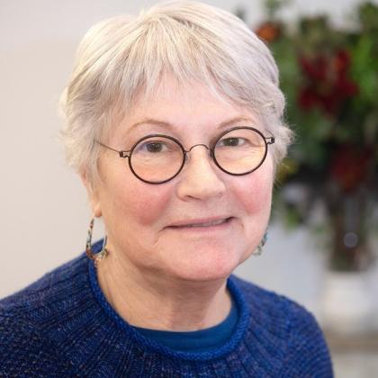 A smiling headshot of Royal Marsden patient, Betsy. She has short, cropped silver hair and round, dark-framed glasses. She's wearing a dark blue knitted jumper and some dangly earrings.