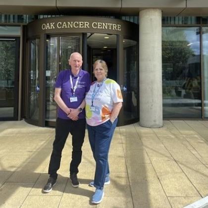 Volunteers Brian and Pat standing outside the entrance to The Royal Marsden's Oak Cancer Centre. They are both wearing lanyards and branded volunteering t-shirts