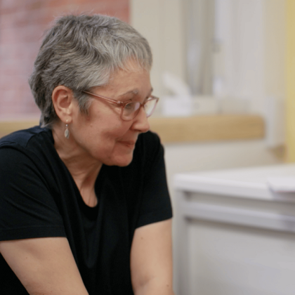 Royal Marsden patient Tracy smiling and shrugging her shoulders while speaking to a surgeon. She has short, cropped grey hair and is wearing a black t-shirt, pink framed glasses and some dangly earrings.