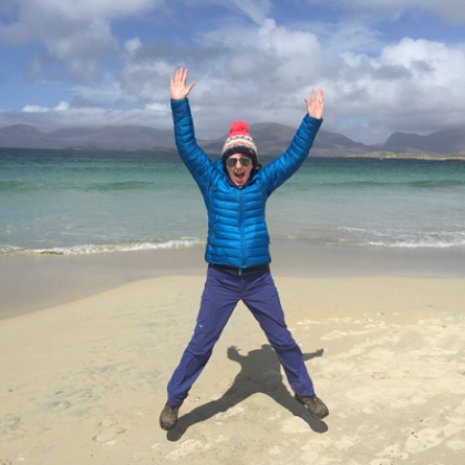 Shiona in hiking gear doing a star jump on a sandy beach. There are mountains in the background  