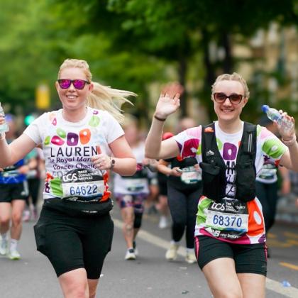 Team Marsden runners, Lauren and Emma, are both smiling and waving at the camera during London Marathon 2025