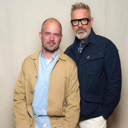Jack Dyson and Daniel Marks, founders of father and son day. Jack is sitting down and wearing a light blue shirt with a cream-coloured jacket over the top. Daniel is standing next to him, wearing dark framed glasses and a navy blue jacket. 
