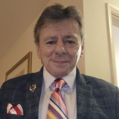 Royal Marsden patient, Steve. He has short, grey hair and is wearing a dark grey suit jacket, with a pale shirt, brooch and colourful-striped tie.