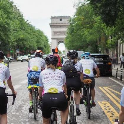 Team Marsden cyclists on London to Paris Cycle, cycling on street in Paris with trees and Arc de Triomphe in the background 