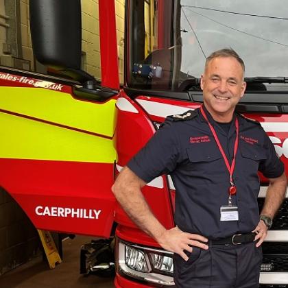 A man in a fire fighter's uniform, standing and smiling in front of a fire engine with his hands on his hips.