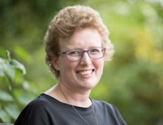 Lizzie smiling. She is wearing a grey t-shirt, square frame glasses and has short curly hair