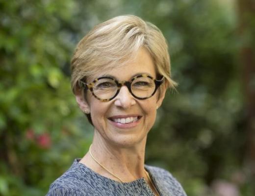 Portrait of Brenda smiling. She has short blonde hair and is wearing round rim glasses