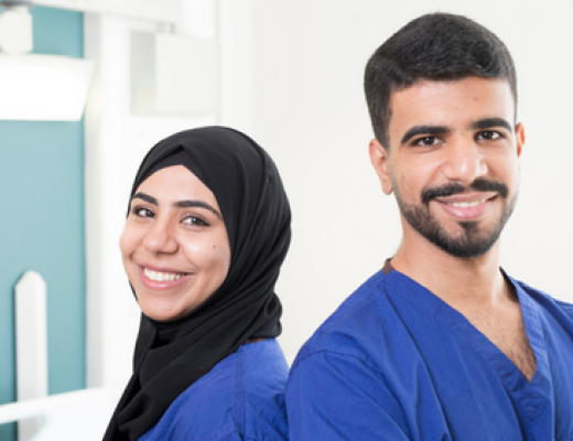 Sara (Left) and Abed (right) wearing blue scrubs and standing back to back. 