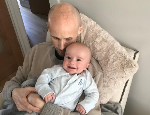 Simon sitting in a arm chair holding baby Dexter who is smiling.