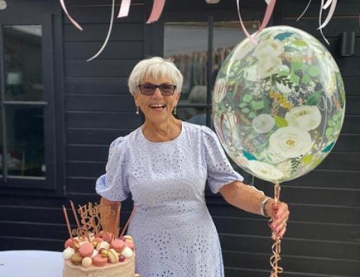 Photo of Jacky, on her 80th birthday she is wearing sunglasses, a white dress and holding a floral balloon. She has a big smile. 
