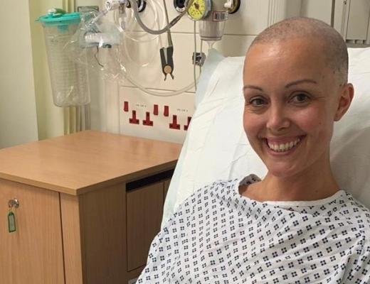 Natalie Hall at The Royal Marsden. She is in a ward and wearing a hospital gown. She has a big smile. 