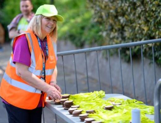 Smiling woman handing out medals. She is wearing a neon orange Banham Marsden March volunteers vest and cap. 