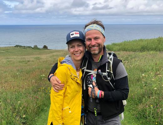 Young man and woman arm in arm on the South downs. They are wearing hiking gear and smiling at the camera. The man has a Royal Marsden Cancer Charity shirt on and the woman wears a bright yellow jacket.
