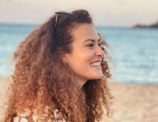 A portrait photo of a woman smiling. She has brown curly hair and is on a beach 