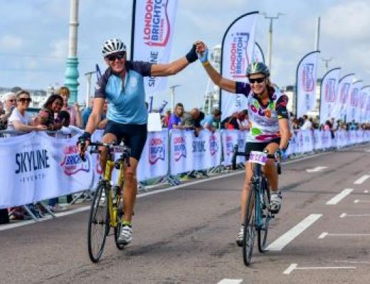 A couple cycling over the London to Brighton Cycle Ride finish line. The are holding hands in the air in celebration and wearing Royal Marsden Cancer Charity cycling vests