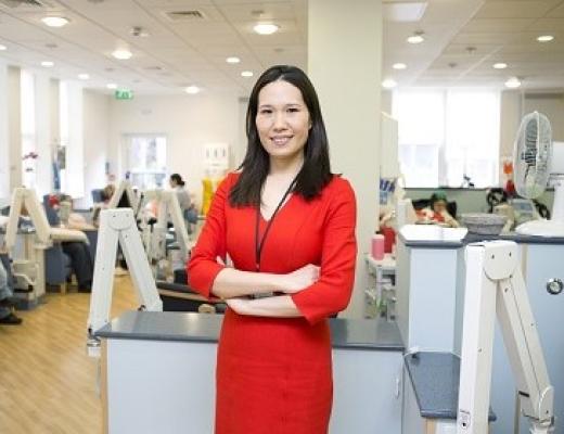 Dr Chong in a smart red dress standing with her arms crossed in a busy hospital setting