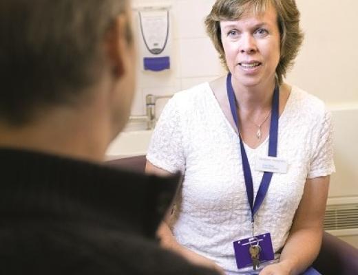 Clare sitting in a hospital consultation room holding a pen and folder, talking to a patient