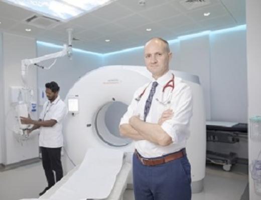 Dr Lee standing in front of a CT Scanner and a radiographer behind him using the machine