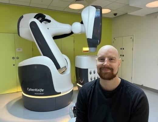  Alastair smiling and standing in front of a large white machine that resembles a robotic arm. 