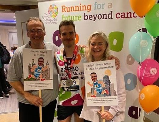 George standing in the middle of his two parents, wearing his running gear and a marathon medal around his neck. They are in front of a Royal Marsden Cancer Charity roll-up banner and balloons and his parents are holding marathon support signs