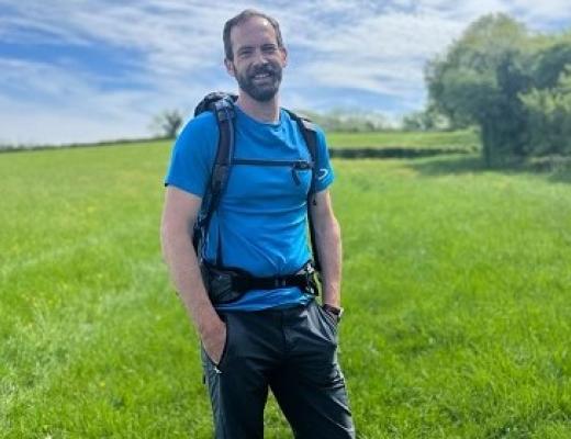 Jon standing in a rolling green field on a sunny day, wearing a backpack and walking gear.