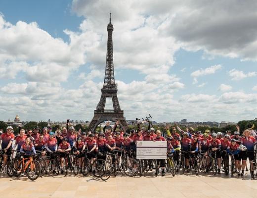  A large group of cyclists dismounted from their bikes, standing and waving in front of the Eiffel tower