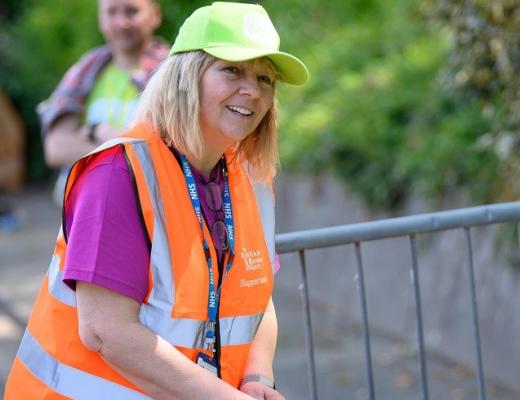 A woman smiling. She is wearing a Banham Marsden March volunteer vest and cap and by a table of finishers medals. 